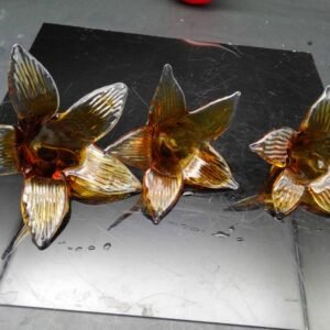 A trio of intricately handcrafted glass flower-shaped chandelier parts, showcasing a rich amber hue with ribbed textures on the petals. Each piece features unique organic curves and subtle imperfections, reflecting the artisanal quality of glassblowing techniques. The flowers are set against a reflective black surface, enhancing their translucent beauty and capturing light playfully.