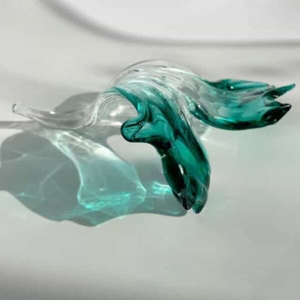 Handcrafted Teal and Clear Glass Sculpture - Elegant Artistry A graceful glass sculpture showcasing flowing lines with a blend of clear and teal glass. The piece appears to undulate like water, highlighting the artisanal craftsmanship and delicate curves typical of hand-blown glass art. The light refracts through the transparent sections, creating a stunning play of shadows on the surface beneath.