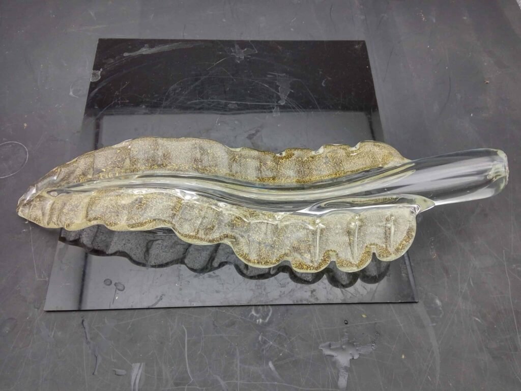 A beautifully handcrafted glass chandelier part featuring a wavy, leaf-like design. The piece showcases delicate textures with irregular surface striations and a warm, translucent golden hue. Hints of tiny air bubbles caught in the glass highlight its handmade nature, differentiating it from machine-produced items. This decorative glass element can enhance various lighting fixtures with its artistic flair.