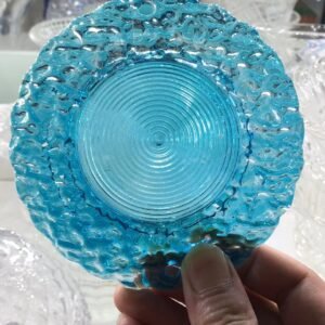 A close-up view of a handcrafted glass piece, showcasing its vibrant blue color and intricate texture. The dish has a wavy edge with organic curves, complemented by circular ribbed patterns in the center. A hand holds the glass, revealing the brilliance of the transparent material in a well-lit environment, with other glassware blurred in the background. This unique glassware exhibits the beauty of artisan craftsmanship, typical of decorative glass chandelier parts or pressed glass designs.